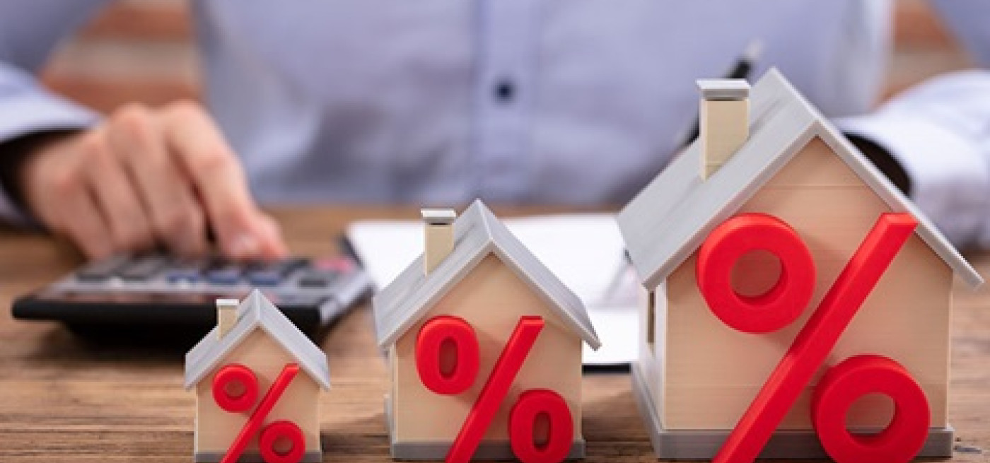 Três maquetes de casas em tamanhos crescentes com símbolos percentuais vermelhos à frente, sobre uma mesa, enquanto uma pessoa utiliza uma calculadora ao fundo, representando taxas de juro e spread no crédito habitação.